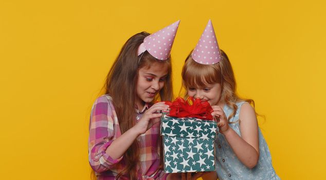 Happy toddler children sisters girls siblings friends wearing festive cone cap celebrating birthday anniversary party, holding gift box. Schoolkids opening present advertising sale discount holidays