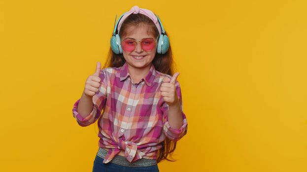 Happy preteen child girl kid listening music via headphones and dancing disco fooling around having fun expressive gesticulating hands. Little toddler children isolated on studio yellow background