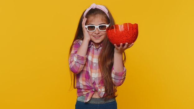 Little toddler children girl in shirt eating popcorn, watching comedy movie film wearing 3D glasses, enjoying cinema snacks. Young preschool kid in glasses isolated alone on yellow studio background