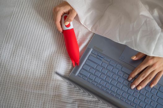 A woman lies in bed holding a clitoral vibrator and watching porn on a laptop. The girl has sex online.