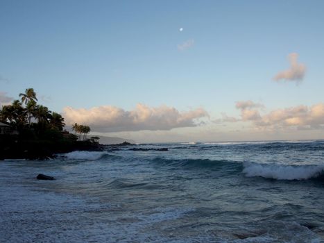 Beautiful Dawn over the ocean with waves crashing into rocks along beach with moon in the sky on the North Shore of Oahu.    