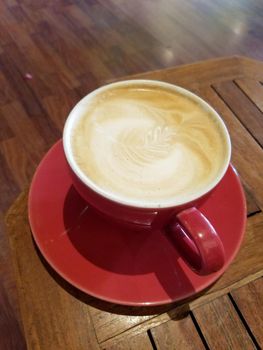 Red cup of Cappuccino on saucer with a leaf pattern in foam on table.