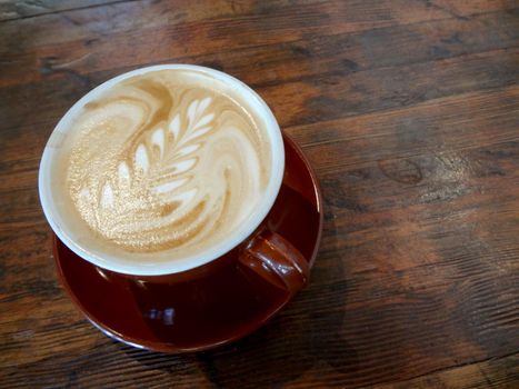 Red cup of Cappuccino on saucer with a leaf pattern in foam on table.