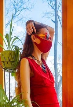 A girl in a red dress, a red medical mask and funny sunglasses straightens her hair