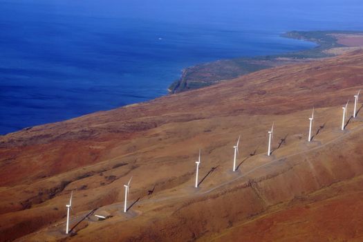 Aerial view of Modern Windmills spin on hillside with windy road and coastline on the island of Maui.