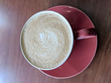 Red cup of Cappuccino on saucer with a leaf pattern in foam on table.