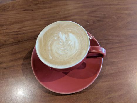 Red cup of Cappuccino on saucer with a leaf pattern in foam on table.