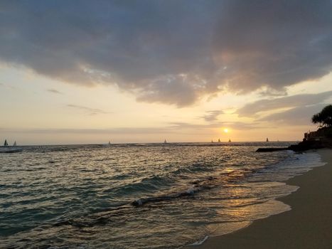 Sunsets over Waikiki waters as waves roll into shore at Makalei Beach Park with sail boats in the ocean on Oahu, Hawaii.