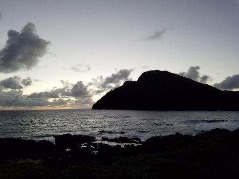 Ocean waves lap on Makapuu beach before dawn with Makapuu Lighthouse and point in the distance looking out to sea on the east side clouds in sky of Oahu, Hawaii.