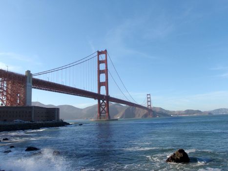 Wave breaks on shore with Golden Gate Bridge, Marin County and Fort Point in distance in San Francisco, California.