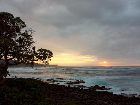 SUP Paddler ride waves as sunsets though the clouds off the coast of Turtle Bay on the North Shore of Oahu, Hawaii.
