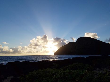 Ocean waves lap on Makapuu beach as sunrises through the clouds with Makapuu Lighthouse and point in the distance looking out to sea on the east side clouds in sky of Oahu, Hawaii.