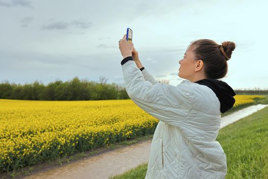 slender and beautiful girl shoots a video of a yellow rapeseed field on her phone. The girl is resting in nature and admiring the bewitching landscape