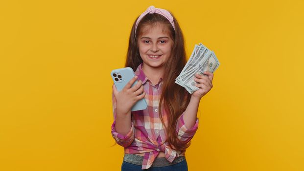 Smiling happy young child girl kid looking smartphone display sincerely rejoicing win, receiving money dollar cash banknotes, success lottery jackpot luck. Little toddler children on yellow background