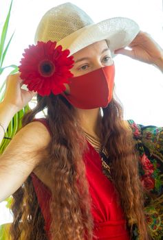 A girl in a red dress, a white hat, funny sunglasses and a red medical mask stands in a floral scarf with plants and a red gerbera against the sky in early spring