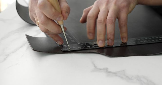 Working process of crafting a new handmade leather wallet in the leather workshop. Man's hands holding awl and iron ruler