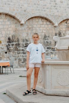 Girl Tourist Resting in the Ancient Narrow Street On A Beautiful Summer Day In MEDITERRANEAN MEDIEVAL CITY, OLD TOWN KOTOR, MONTENEGRO. Young Beautiful Cheerful Woman Walking On Old Street