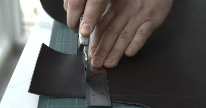 Working process of crafting a new handmade leather wallet in the leather workshop. Man's hands working with knife and iron ruler and cutting a leather