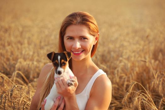 Young woman smiling, holding Jack Russell terrier puppy, sunset lit wheat field in background.