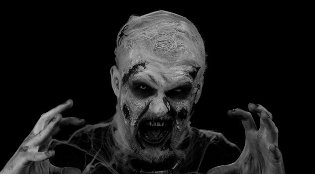 Creepy man with Halloween zombie bloody wounded makeup, trying to scare, face expressions. Horror theme. Sinister undead guy isolated on studio black background. Voodoo rituals. Fashion body art