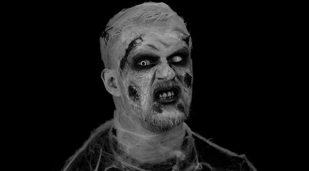 Frightening man with Halloween zombie bloody wounded makeup, trying to scare, face expressions. Horror theme. Sinister undead guy isolated on studio black background. Voodoo rituals. Fashion body art