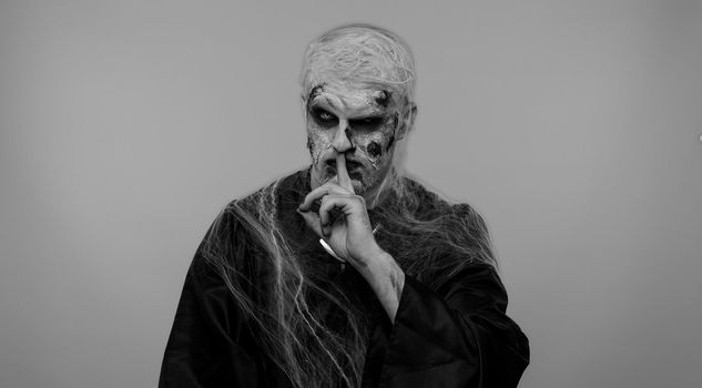 Shh be quiet. Sinister man with horrible scary Halloween zombie makeup in costume presses index finger to lips makes silence gesture sign do not tells secret. Dead guy with wounded bloody scars face
