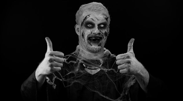 Ugly man with scary Halloween zombie makeup in costume raises thumbs up agrees with something or gives positive reply recommends advertisement likes good. Dead guy with wounded bloody scars face