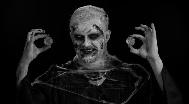Zombie man with make-up with fake wounds scars showing golden bitcoins. Achievement career wealth, cryptocurrency investment, mining btc, future technology. Sinister dead guy. Halloween, filming
