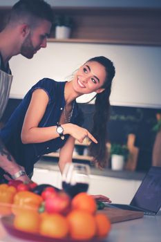 Young man cutting vegetables and woman standing with laptop in the kitchen.