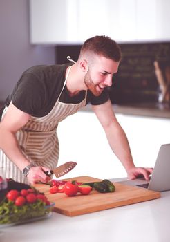 Portrait of man cooking vegetable in the kitchen while looking at a laptop computer on the table.