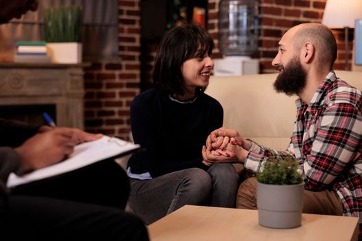 Happy couple having romantic moment holding hands at therapy session with psychologist, solving relationship difficulties and enjoying reconciliation. Receiving counseling advice and help.