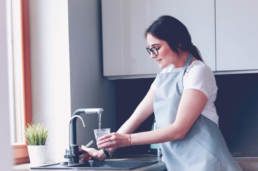 Human hand holding glass pouring fresh drink water at kitchen faucet.