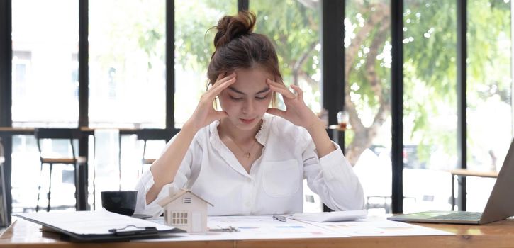 Photo of a tired woman keeps a hand on her head at the wooden working desk after finishing work hard..