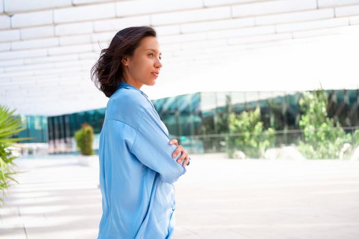 Side view Confidence Businesswoman portrait with crossed hands. Pretty business woman 30 years old standing near office building dressed blue shirt. Caucasian female business person outside