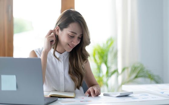 Beautiful Asian businesswoman analyzes charts using laptop calculator at the office..