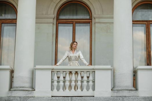 a beautiful mature woman in a long white dress stands on the balcony and looks into the distance.