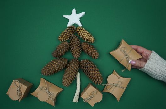 On a green background, a woman's hand laid out gifts in craft paper and made a Christmas tree out of fir cones. High quality photo