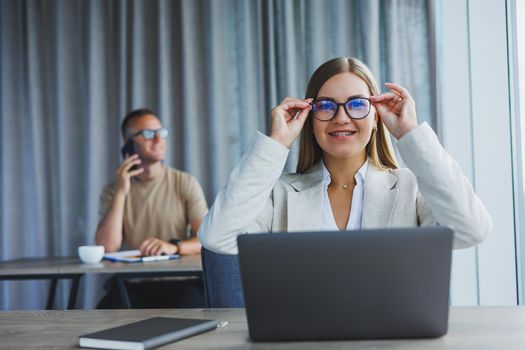 Attractive young woman in glasses sits at a table with a laptop in a coworking space and discusses a project plan with a colleague. Workflow in the office.