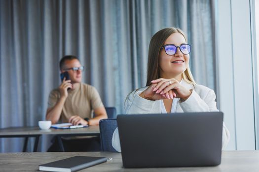 Attractive young woman in glasses sits at a table with a laptop in a coworking space and discusses a project plan with a colleague. Workflow in the office.