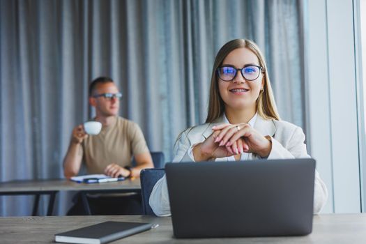 Attractive young woman in glasses sits at a table with a laptop in a coworking space and discusses a project plan with a colleague. Workflow in the office.