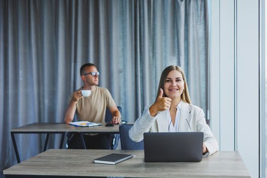 Attractive young woman in glasses sits at a table with a laptop in a coworking space and discusses a project plan with a colleague. Workflow in the office.