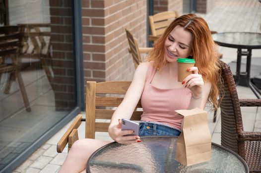 Young red-haired woman with braces on her teeth drinks coffee on a summer veranda