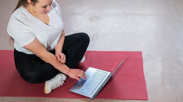 A chubby young woman is watching an online fitness lesson on a laptop. Distance sports training