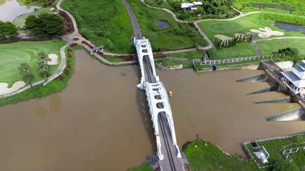 Aerial view of Tha Chomphu White Bridge, Lamphun, Thailand with river, forest trees and green mountain hill. An old railway bridge across the river. Tourist attraction landmark.