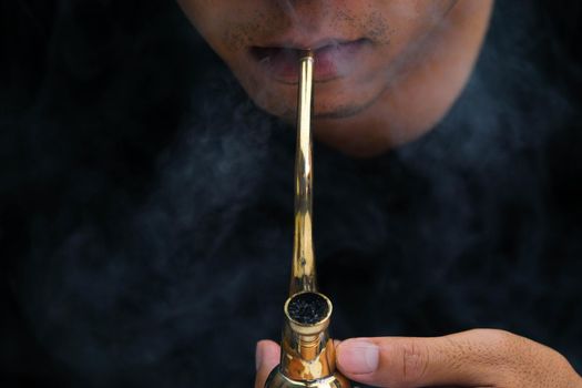 Asian man smokes marijuana from a pipe at home. Studio shoot with model simulating smoking pot with a pipe in a dark background. Cannabis legalisation.