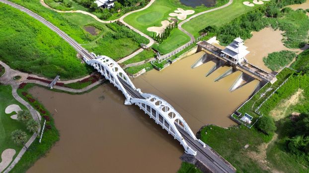 Aerial view of Tha Chomphu White Bridge, Lamphun, Thailand with river, forest trees and green mountain hill. An old railway bridge across the river. Tourist attraction landmark.