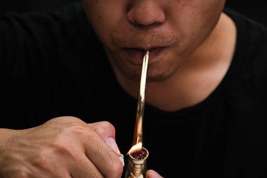 Asian man smokes marijuana from a pipe at home. Studio shoot with model simulating smoking pot with a pipe in a dark background. Cannabis legalisation.