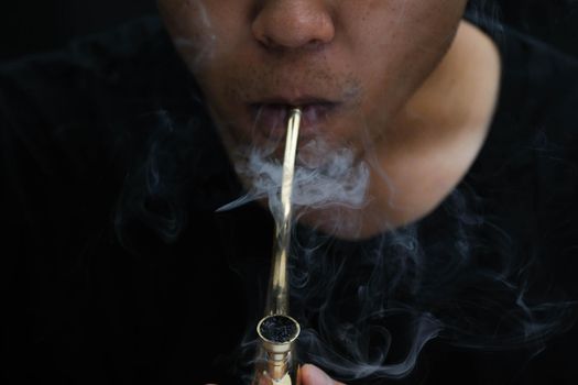 Asian man smokes marijuana from a pipe at home. Studio shoot with model simulating smoking pot with a pipe in a dark background. Cannabis legalisation.