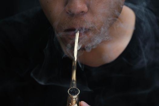 Asian man smokes marijuana from a pipe at home. Studio shoot with model simulating smoking pot with a pipe in a dark background. Cannabis legalisation.