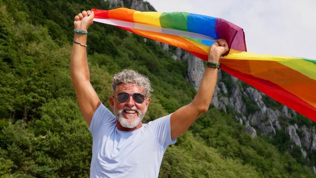 Portrait of a gray-haired senior elderly Caucasian man bisexuality with a beard and sunglasses holding a rainbow LGBTQIA flag on nature. Celebrates Pride Month, Rainbow Flag Day, gay parade